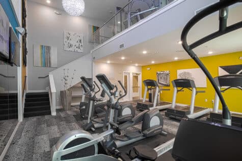 Spacious two-level apartment gym featuring cardio equipment including treadmills, ellipticals, and rowing machines. The room includes a staircase to an upper mezzanine, modern artwork, yellow accent walls, and large mirrors for an open, upscale fitness environment.