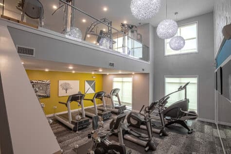 Two-story luxury fitness center with treadmills, ellipticals, and cardio equipment arranged beneath decorative pendant lighting. The open-concept gym includes bright accent walls, large windows, mezzanine-level workout space, and modern contemporary finishes.