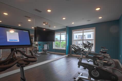 Modern spin and cycling studio with stationary bikes, mirrored walls, mounted TVs, and large windows providing natural light. The room features teal accent walls, dark wood flooring, and a clean, upscale fitness design.