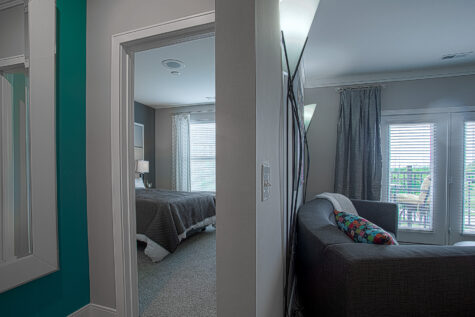 Modern apartment interior at Savoy at the Streets of West Chester in West Chester, Ohio featuring a hallway view connecting the bedroom and living area, with neutral gray finishes, contemporary furnishings, large windows with blinds, and a glimpse of the spacious open-concept layout by HILLS Properties.