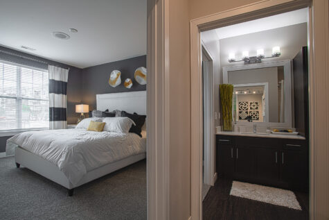 Primary bedroom and bathroom suite at Savoy at The Streets of West Chester featuring a bright bedroom with large windows, plush carpeting, and a contemporary bathroom with dark wood cabinetry, quartz countertops, and modern lighting. The open layout highlights the convenient connection between living and bath spaces.
