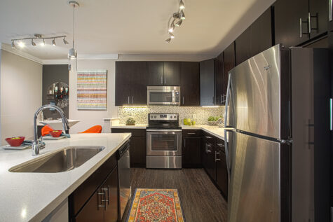 Modern apartment kitchen featuring dark wood cabinetry, stainless steel Whirlpool appliances, quartz countertops, a mosaic tile backsplash, under-cabinet lighting, and a large island with sink at Savoy at the Streets of West Chester.