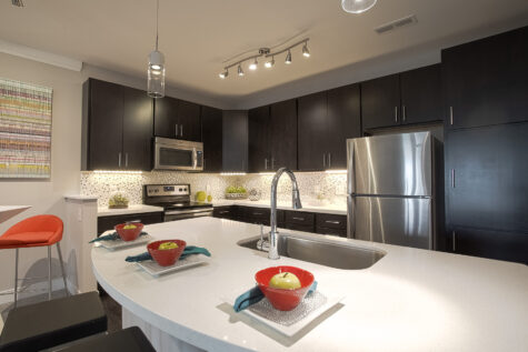 Spacious contemporary kitchen with a large quartz island, stainless steel appliances, dark cabinetry, glass tile backsplash, and pendant lighting showcasing the modern finishes at Savoy at the Streets of West Chester.