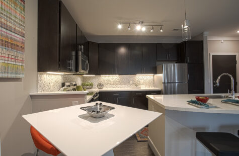 Open-concept apartment kitchen and dining area featuring dark wood cabinets, stainless steel appliances, quartz countertops, island seating, modern lighting, and sleek finishes at Savoy at the Streets of West Chester.