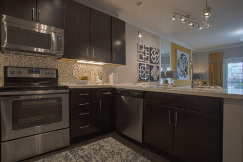 Contemporary kitchen at Savoy at The Streets of West Chester featuring dark espresso cabinetry, stainless steel appliances, quartz countertops, a mosaic tile backsplash, and a breakfast bar with pendant lighting. The open-concept layout flows seamlessly into the living area for modern apartment living.
