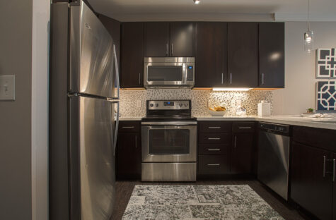 Modern apartment kitchen at Savoy at The Streets of West Chester showcasing stainless steel appliances, dark wood cabinets, quartz countertops, under-cabinet lighting, and a decorative tile backsplash. Ample storage and workspace create a functional and stylish cooking environment.