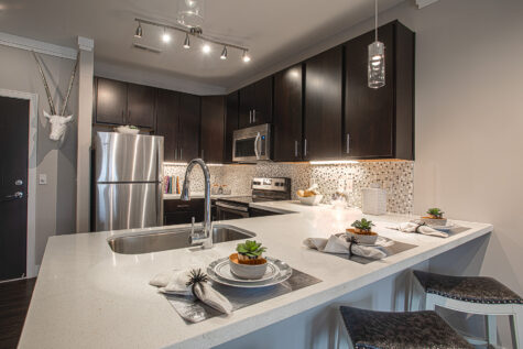 Spacious kitchen with a large quartz island at Savoy at The Streets of West Chester featuring an undermount sink, stainless steel appliances, dark cabinetry, pendant lighting, and bar seating. Contemporary finishes and an open design make the space ideal for cooking, dining, and entertaining.