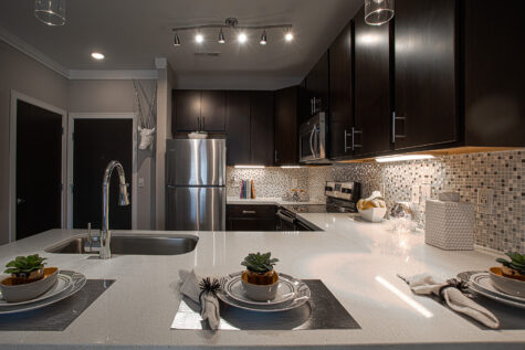 Contemporary kitchen at Savoy at The Streets of West Chester featuring a large quartz island with seating, an undermount sink with gooseneck faucet, dark espresso cabinetry, stainless steel appliances, and a mosaic tile backsplash. Pendant and track lighting illuminate the open-concept space, creating a stylish setting for cooking, dining, and entertaining.