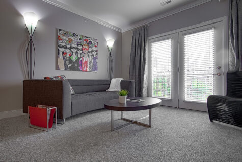 Modern apartment living room featuring plush carpet flooring, a gray sofa, contemporary accent chairs, decorative wall art, floor lamps, and large windows with blinds providing natural light at Savoy at the Streets of West Chester.