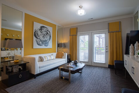 Modern apartment living room at Sterling Lakes in Mason, Ohio featuring a white sofa, dark wood coffee table, large area rug, contemporary artwork, floor-to-ceiling windows with blinds, and stylish black and gold accents.