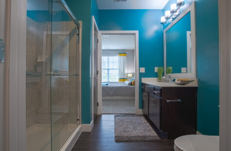 Spacious modern bathroom featuring a glass walk-in shower, dark wood vanity with quartz countertop, large mirror, teal accent walls, and views into the adjoining bedroom area at Savoy at the Streets of West Chester.