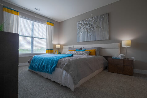 Modern apartment bedroom featuring a spacious layout, plush carpet flooring, a large upholstered bed, contemporary nightstands with lamps, decorative wall art, and a large window with blinds providing natural light at Savoy at the Streets of West Chester.
