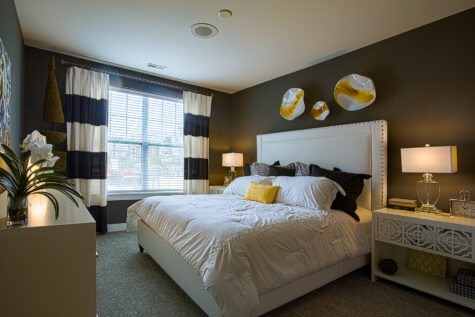 Elegant apartment bedroom at Sterling Lakes in Mason, Ohio featuring a large upholstered bed with white bedding, dark accent wall, modern bedside tables with lamps, decorative wall art, and a spacious window with curtains allowing natural light.