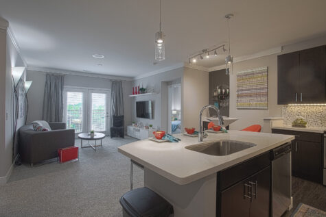 Open-concept apartment interior featuring a modern kitchen with quartz countertops, dark cabinetry, stainless steel appliances, island seating, and a spacious living area with natural light at Savoy at the Streets of West Chester.