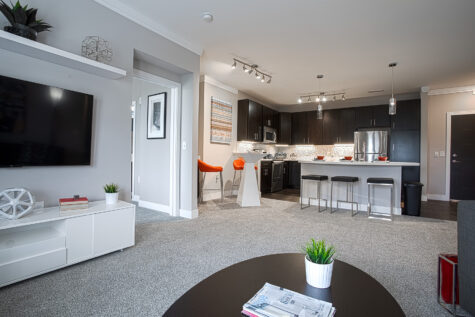 Spacious open-concept apartment featuring a modern kitchen with dark cabinetry, stainless steel appliances, quartz countertops, island seating, and a connected living area with a wall-mounted TV and contemporary finishes at Savoy at the Streets of West Chester.