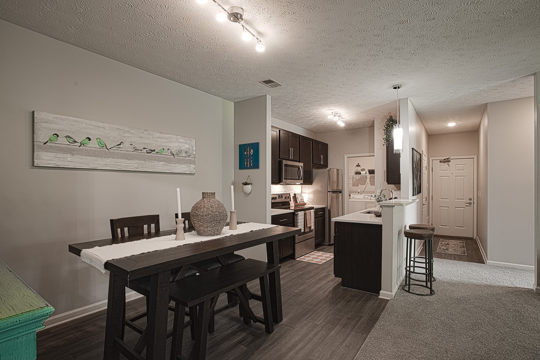 Open-concept apartment interior at Sterling Lakes in Mason, Ohio, featuring a dining area with a dark wood table and bench seating, a modern kitchen with espresso cabinets and stainless steel appliances, a breakfast bar with seating, and a bright entryway with neutral finishes.
