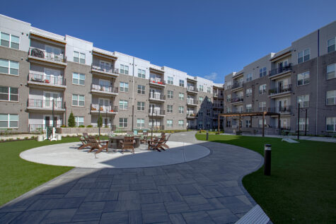 Expansive resident courtyard at Vantage Anderson by HILLS Properties in Anderson, Ohio, featuring a central fire pit, outdoor grilling pavilion, landscaped walking paths, and spacious green space surrounded by modern apartment buildings.