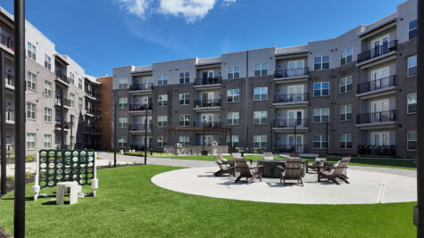Landscaped resident courtyard at Vantage Anderson by HILLS Properties in Anderson, Ohio, featuring a community fire pit with Adirondack seating, oversized Connect Four game, outdoor grilling pavilion, and spacious green space surrounded by modern apartment buildings.