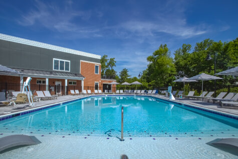 Luxury swimming pool at Relay 129 Apartments and Townhomes showcasing a tanning ledge with in-water loungers, spacious pool deck seating, and a modern clubhouse backdrop.