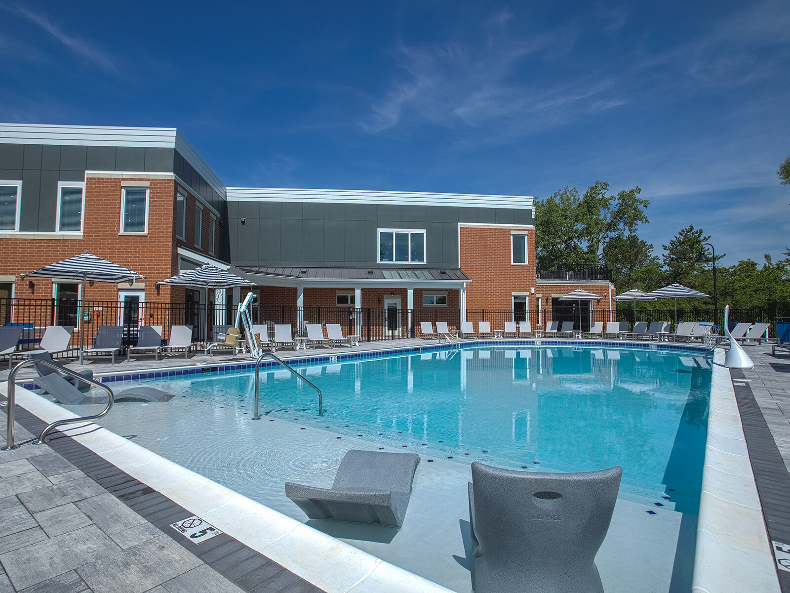 Resort-style swimming pool at Relay 129 Apartments and Townhomes in Liberty Township, Ohio, featuring an expansive sundeck, in-water lounge seating, shaded umbrellas, and scenic wooded views.