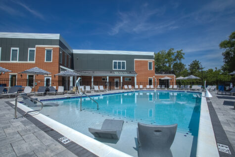 Resort-style swimming pool at Relay 129 Apartments and Townhomes in Liberty Township, Ohio, featuring an expansive sundeck, in-water lounge seating, shaded umbrellas, and scenic wooded views.