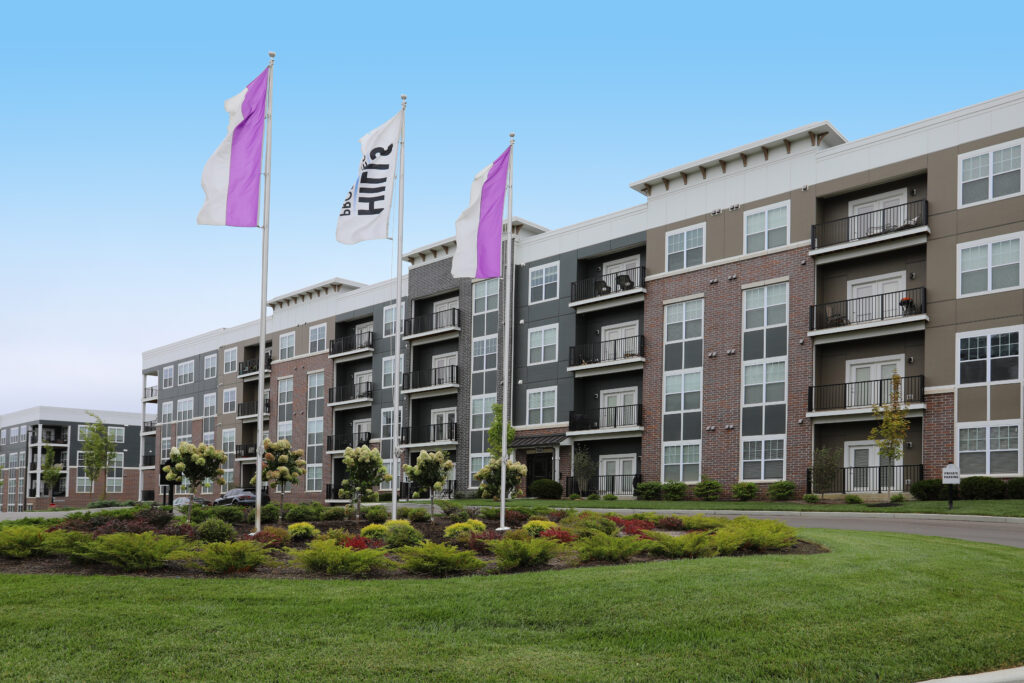 Exterior of a four-story Allure apartment community with brick and mixed-tone façades, private balconies, and white and purple branded flags out front, framed by manicured landscaping and a green lawn.